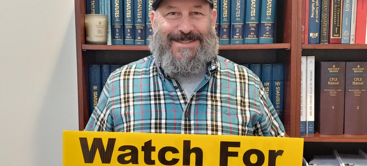 Marketing guy Chris Genovese stands while holding an ABATE "Watch for Motorcycles" lawn sign in front of a bookshelf of legal texts
