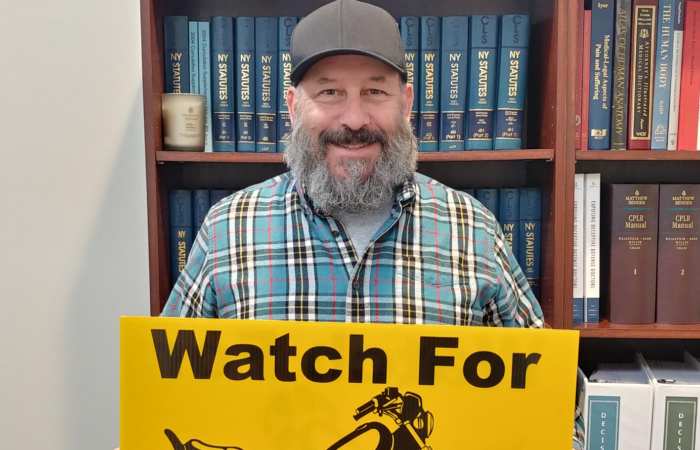 Marketing guy Chris Genovese stands while holding an ABATE "Watch for Motorcycles" lawn sign in front of a bookshelf of legal texts