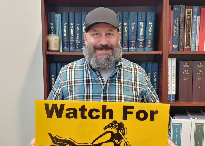 Marketing guy Chris Genovese stands while holding an ABATE "Watch for Motorcycles" lawn sign in front of a bookshelf of legal texts