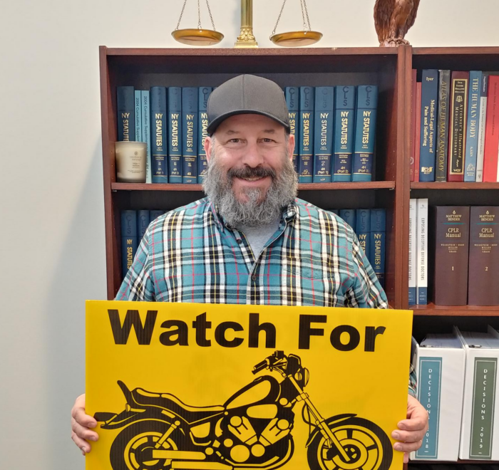 Marketing guy Chris Genovese stands while holding an ABATE "Watch for Motorcycles" lawn sign in front of a bookshelf of legal texts