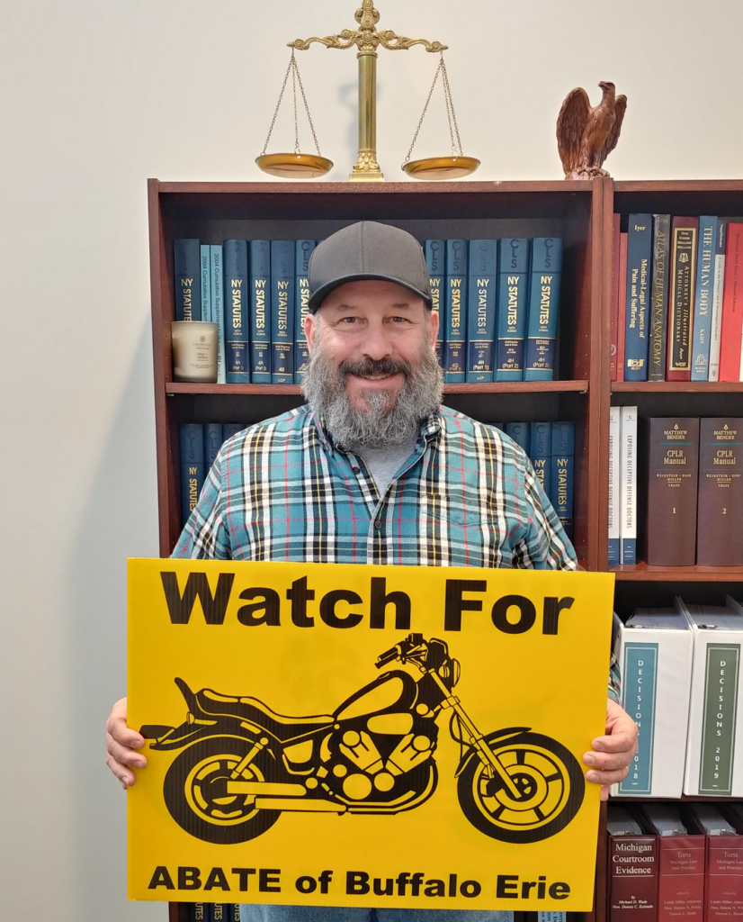 Marketing guy Chris Genovese stands while holding an ABATE "Watch for Motorcycles" lawn sign in front of a bookshelf of legal texts