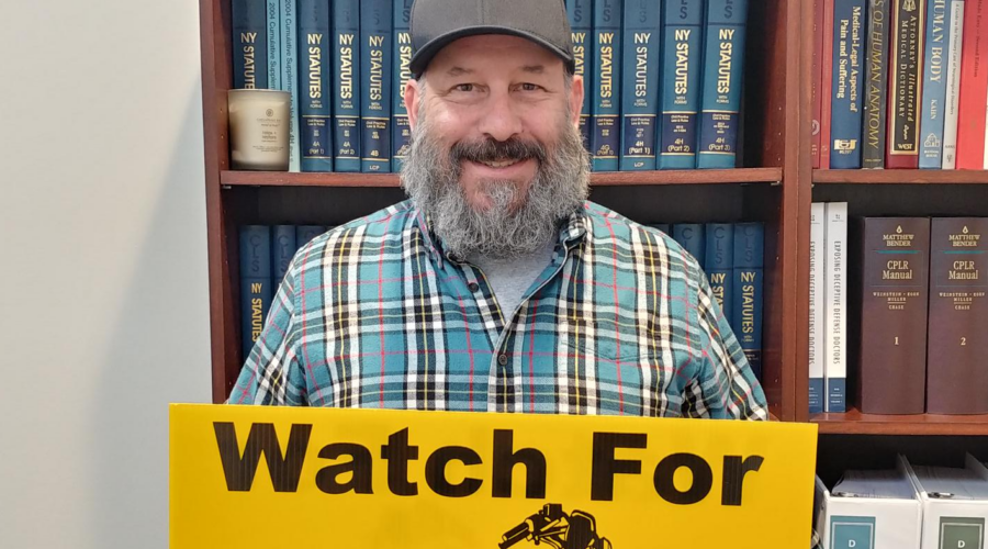 Marketing guy Chris Genovese stands while holding an ABATE "Watch for Motorcycles" lawn sign in front of a bookshelf of legal texts