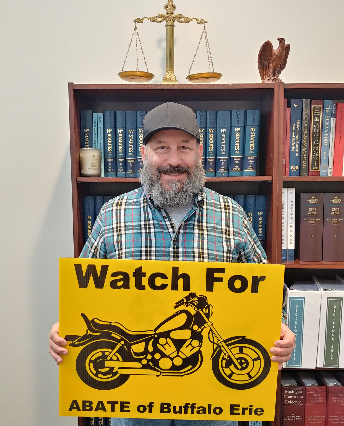 Marketing guy Chris Genovese stands while holding an ABATE "Watch for Motorcycles" lawn sign in front of a bookshelf of legal texts