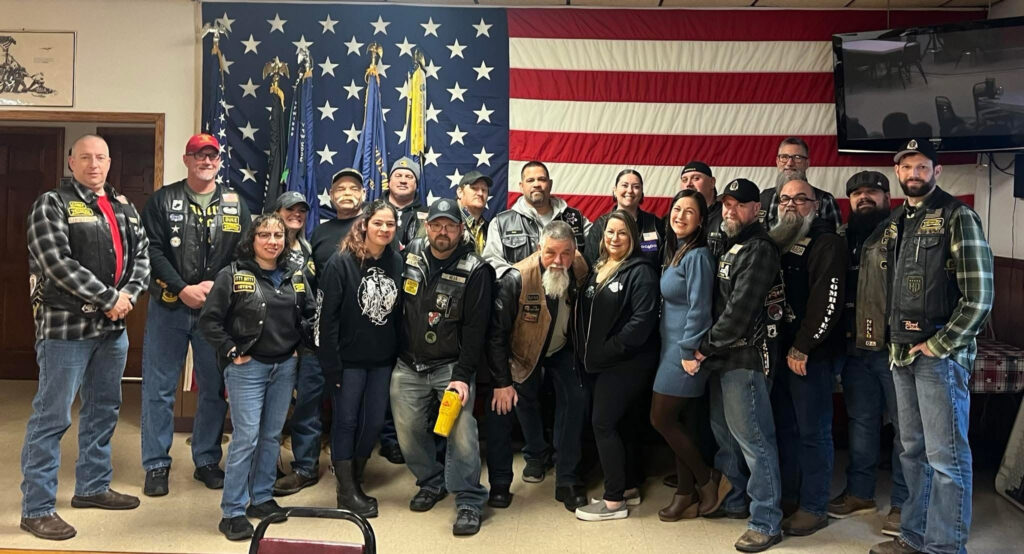 Combat Veterans Motorcycle Association 19-11 members posing for a group photo in front of a large American flag. The photo was taken at a recent chapter meeting. There are about 20 individuals in the phto.
