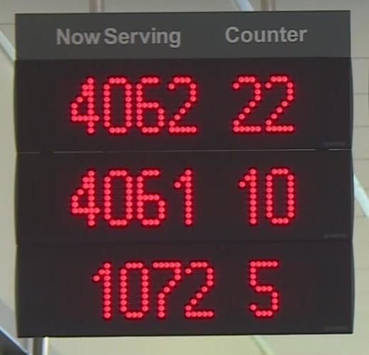 An illuminated "now serving" sign with red lights showing an array of large numbers beneath "now serving" and another array of smaller numbers below "counter." The numbers indicate a large volume of customers and counters where they could be served, most likely indicating a long wait for service.